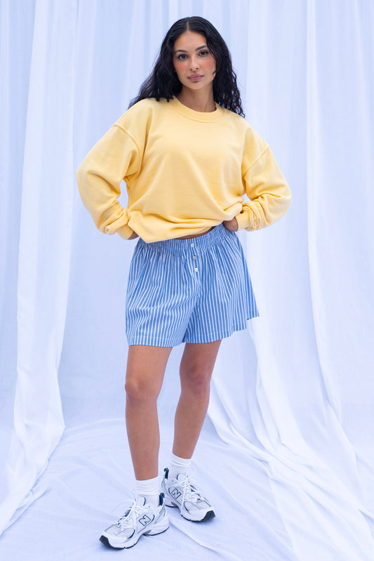 Woman wearing a yellow sweatshirt and blue striped shorts against a white curtain background