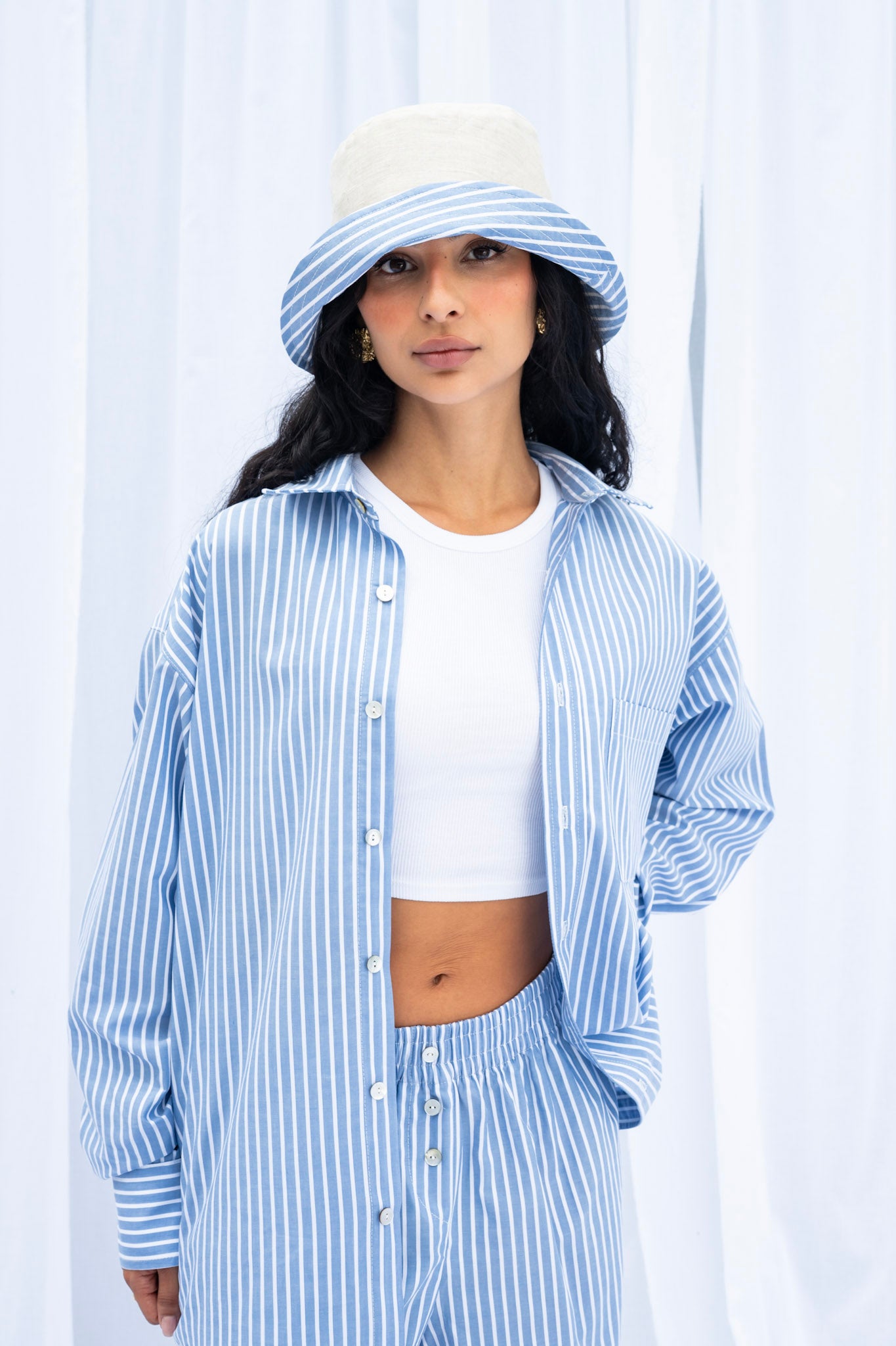Woman wearing a blue and white striped shirt, white top, and matching pants with a white bucket hat against a light background.
