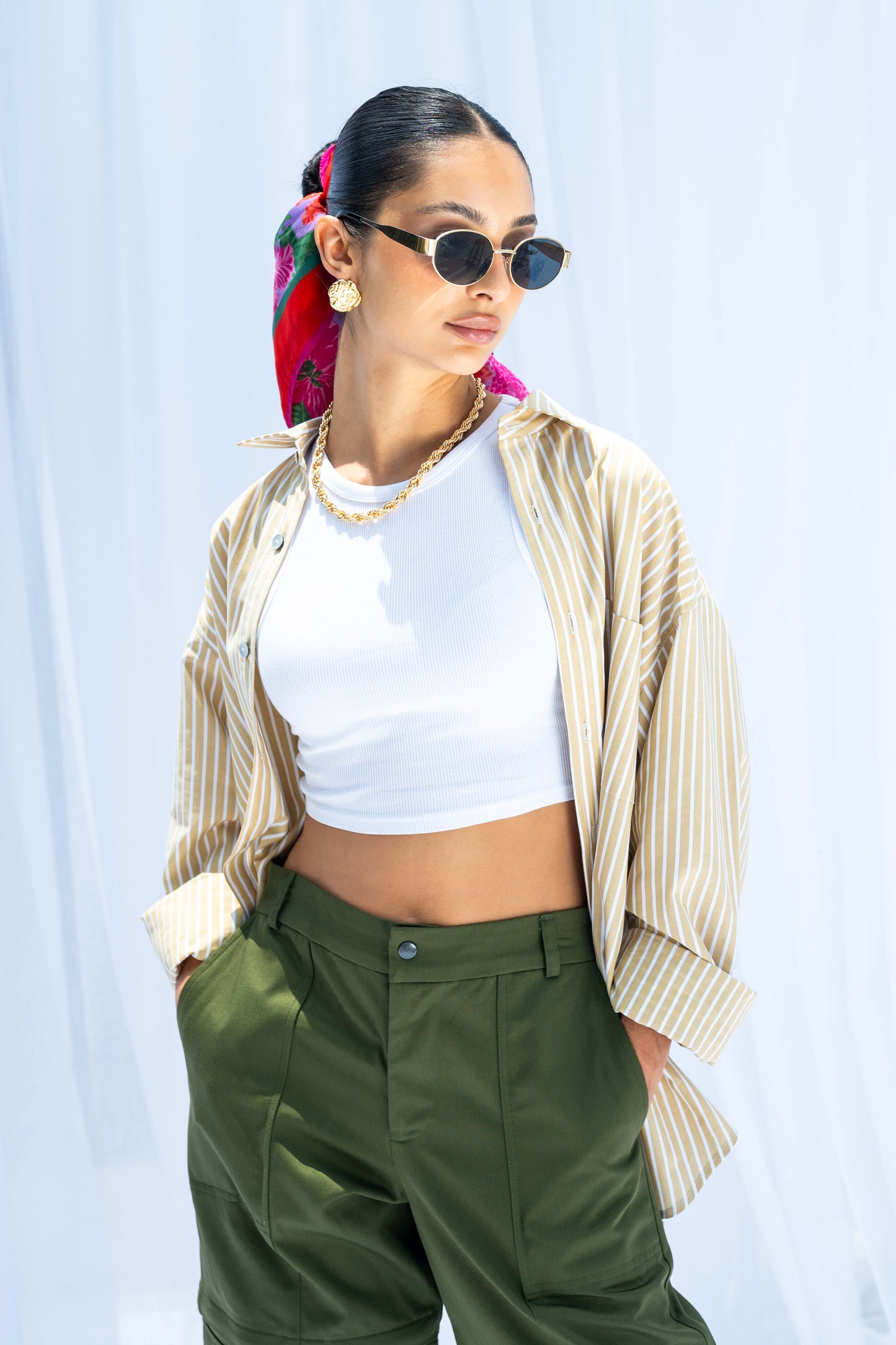 Woman wearing a white crop top, beige striped shirt, green pants, and sunglasses against a light blue background.