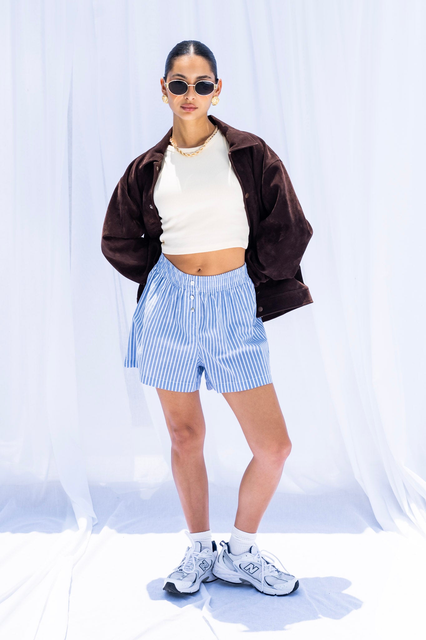Person wearing a brown jacket, white top, blue striped shorts, and white sneakers on a white background