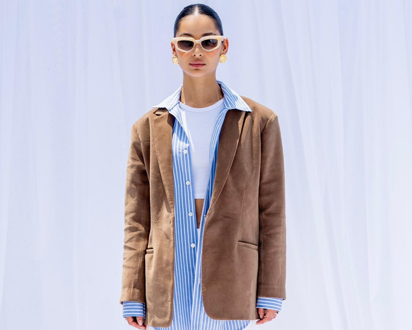 Person wearing a brown blazer, striped shirt, and white pants on a light blue background