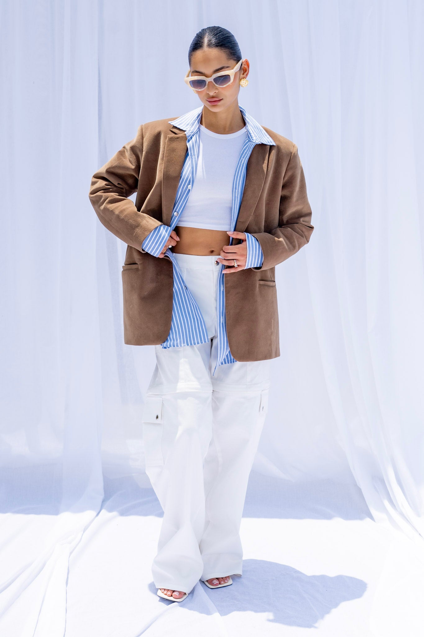 Person wearing a brown jacket, white shirt, and white pants against a light blue curtain background.